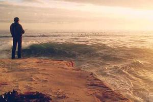 A man stands on the edge of a cliff looking out to a rough sea.